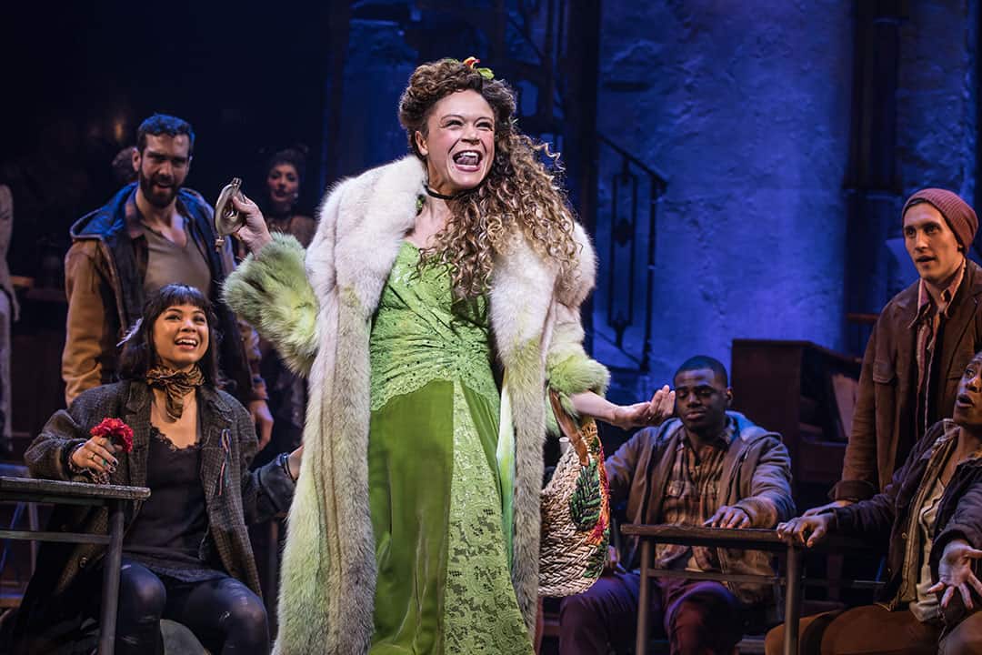 A woman in a long silk dress and elaborate fur coat dances wildly on stage, many of the cast members look on smiling