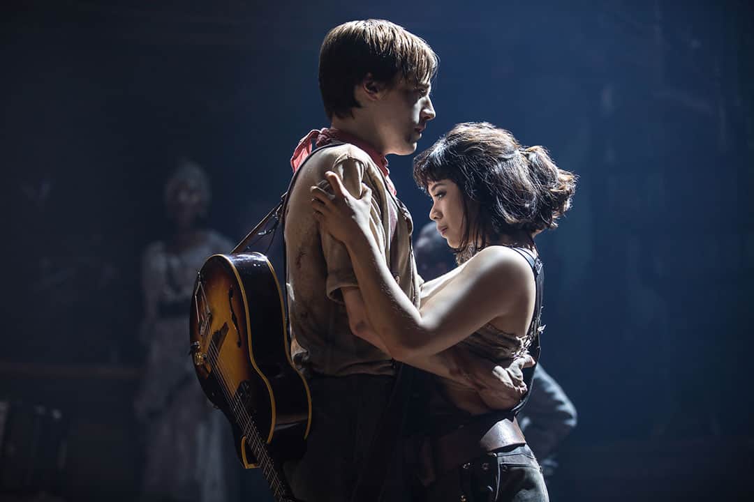 a man with a guitar strapped to his back holds a woman who cannot meet his eyes; they are performers on a stage, backlit and seeming to glow in the stage lights
