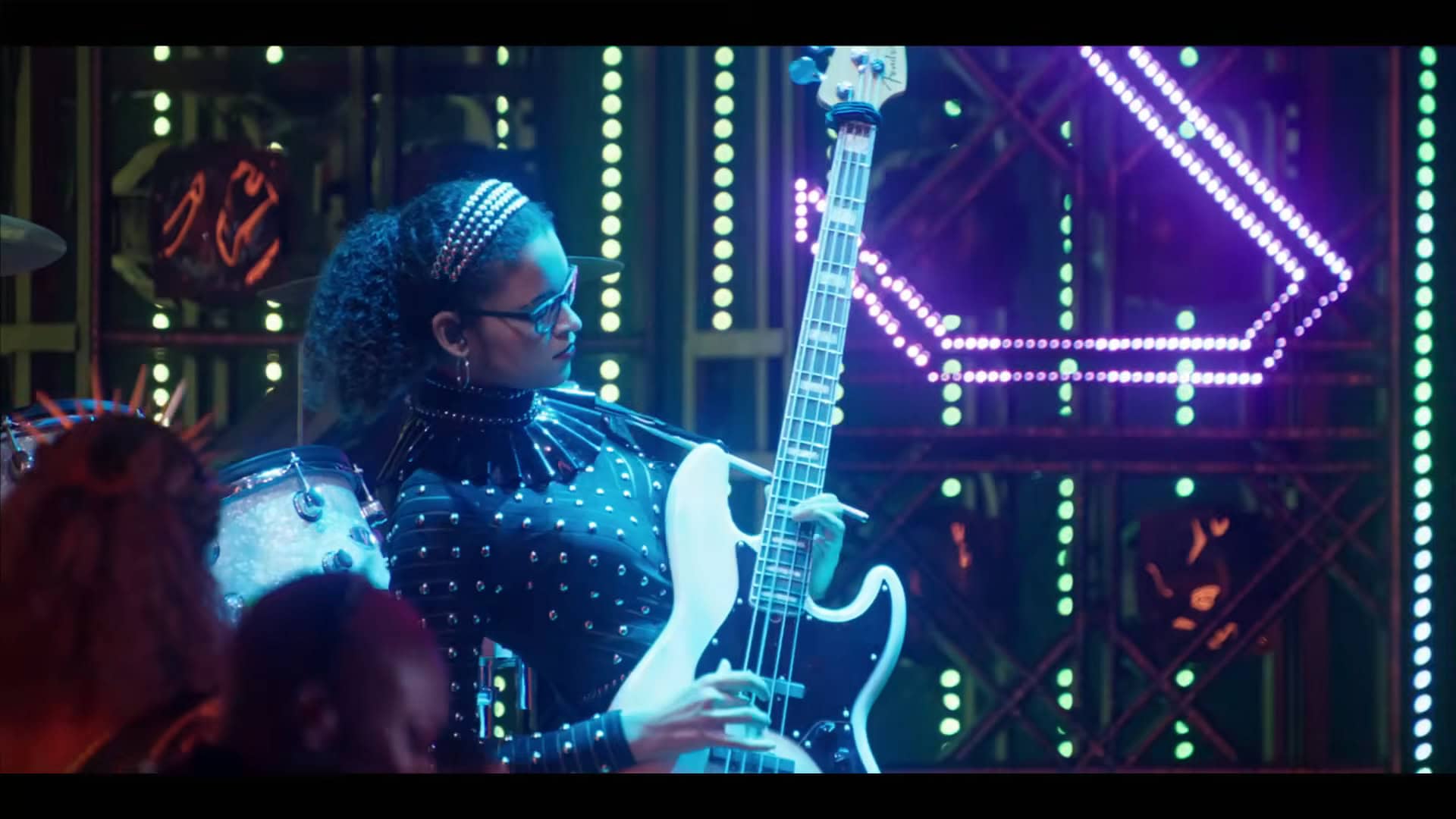A woman wearing glasses with her hair pulled back stands on a stage with a lot of bright lights behind her including a large pink S, She is playing a white bass guitar