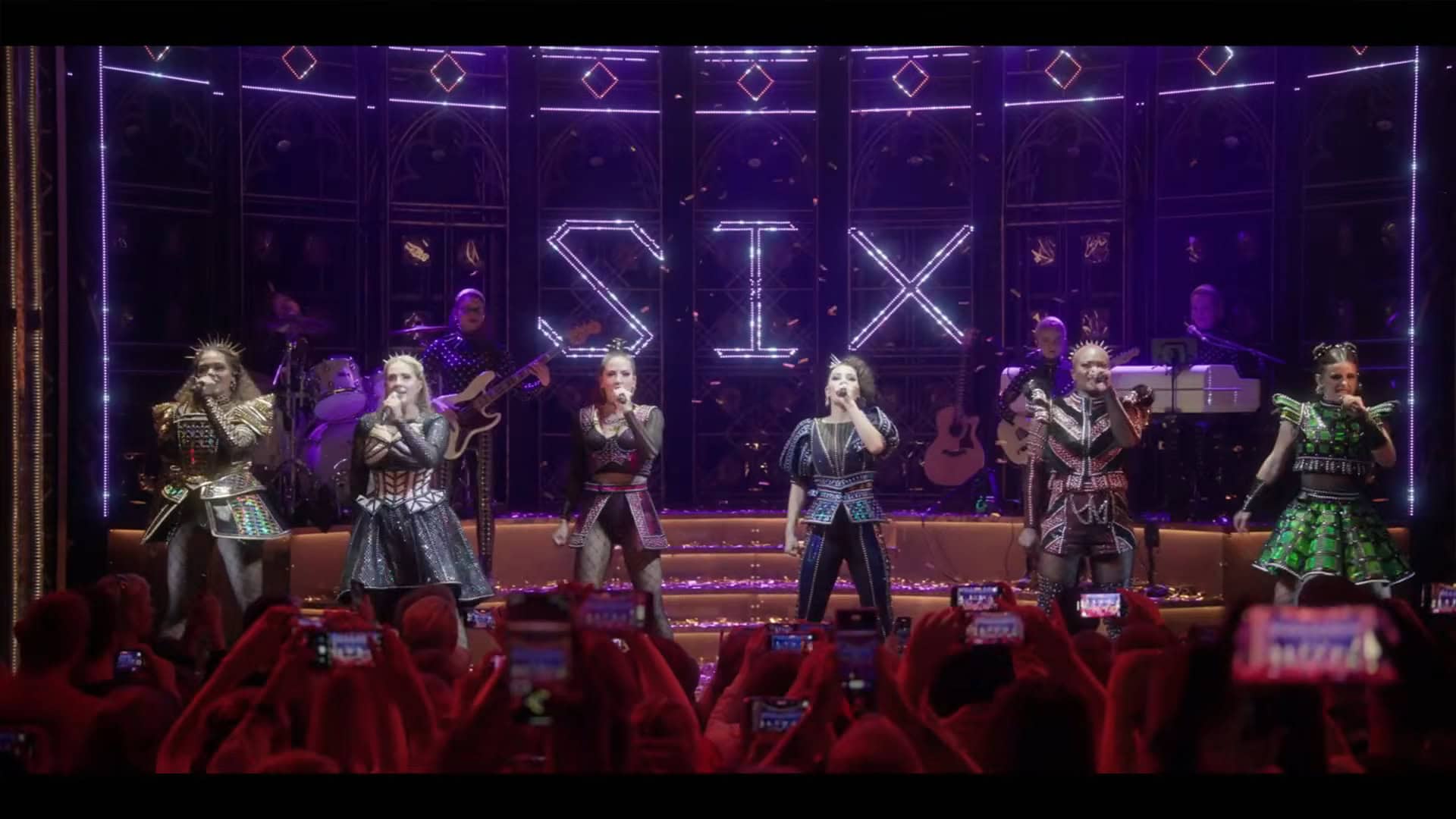 The cast poses in a line spread across the stage for their final number. The band is in the background and the stage is lit brightly. The word "SIX" is spelled in lights across the back of the stage. In the foreground you can see the silhouettes of the heads and hands of some audience members.