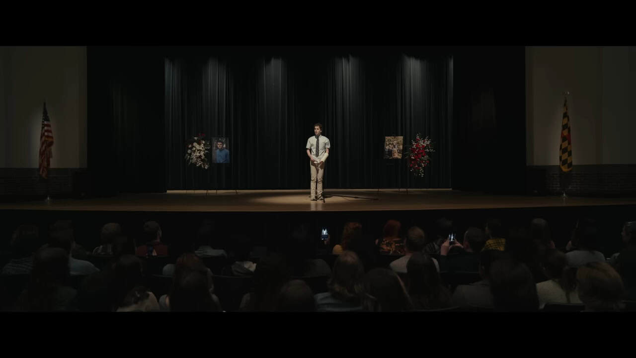 A still from the film Dear Evan Hansen. A teenage boy in slacks, a button up shirt, and tie stands on a stage in a darkened high school auditorium with a spotlight on him, hands clasped, looking nervous. People in the audience gaze up at him, some with their phones out, filming. There are large memorial photos and flowers to either side of him on the stage, similar to a funeral service.