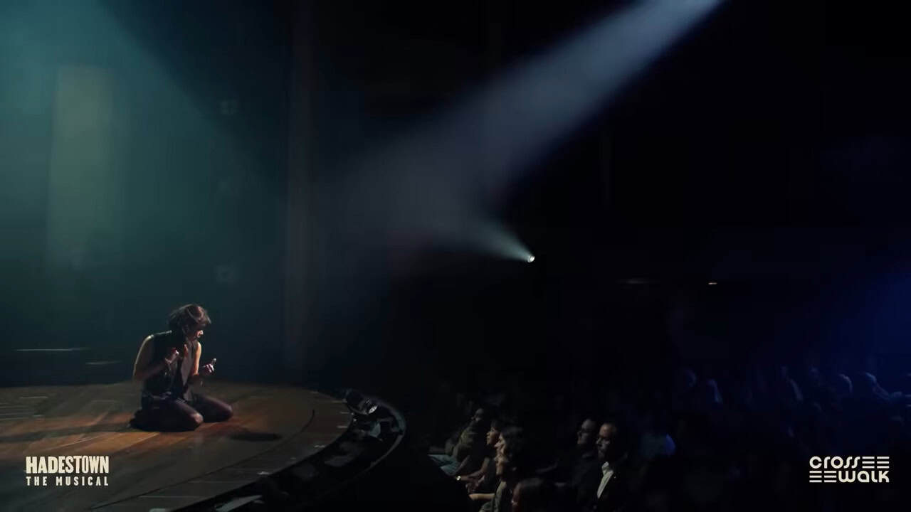 a woman kneels on the edge of the stage in a dark theater with a single spotlight focused on her. We can see the audience gazing up at her in rapt attention