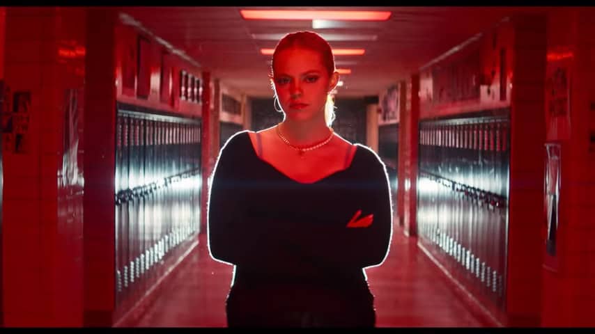 Renee Rapp playing Regina George stands centered in a high school hallway. The lighting is blood red and she is slightly backlit, giving her an otherworldly look as she crosses her arms and stares straight into the camera. This is a scene from the 2024 Mean Girls film adaptation from her song World Burn.