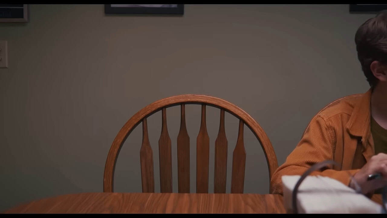 At a kitchen table, an empty chair is centered. The chair next to it has a teenager sitting in it, but we can only see his elbow and the basket of napkins in the center of the table.