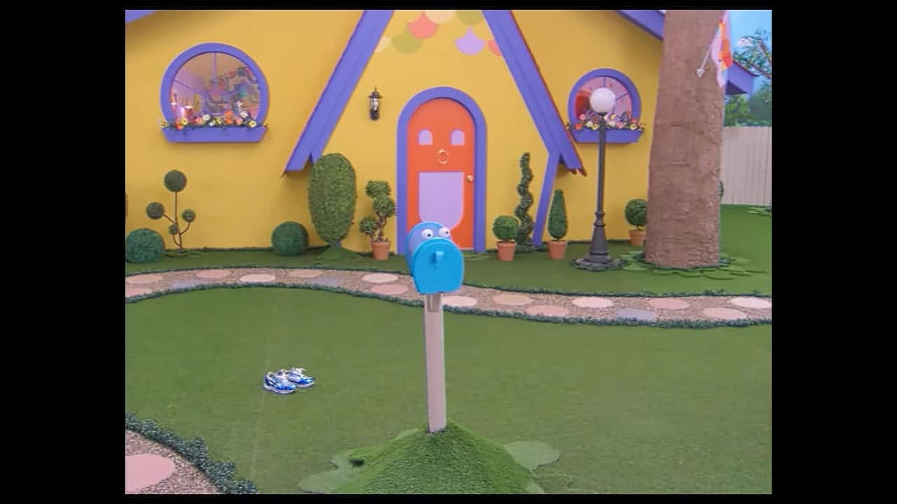 On a television show soundstage, a brightly colored clubhouse sits in the background of an AstroTurf yard, bordered by a cobblestone path and tall fence. The yard is empty except for a blue puppet mailbox with large googly eyes who is watching something off-camera we cannot see and looking upset and two abandoned kids' shoes in the background.