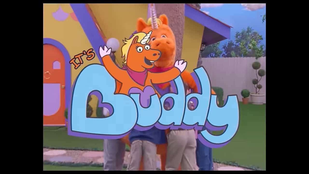 A bright orange unicorn mascot gets a group hug from a group of kids on a TV set. A clubhouse and tall fence are in the background. The logo of the TV show is superimposed over the image reading "It's Buddy"